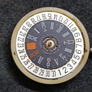 Puede incluir: Un componente de movimiento de reloj circular con una esfera negra con números romanos, un indicador del día de la semana y un anillo exterior blanco que muestra los números del 1 al 31. El texto "O-RING UPWARD" aparece en la esfera.
