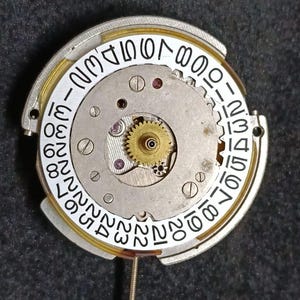 May include: A circular mechanical watch movement component featuring a white date wheel with black numerals from 1 to 31. The centre displays a brass-coloured gear, metallic plates, and small screws, with a thin metal stem extending from the bottom.