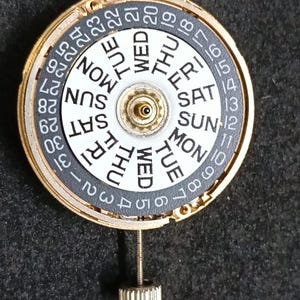 May include: A circular watch movement mechanism featuring a gold-toned outer ring and a central white disc displaying days of the week in black text. A dark grey outer ring shows numbers 1 through 31 in white. A metal winding stem extends from the bottom.