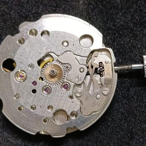 Puede incluir: Un movimiento de reloj mecánico de color plateado que presenta una placa base metálica circular con engranajes visibles, pequeños rubíes sintéticos de color púrpura y una tija de remontuar metálica con una corona estriada.