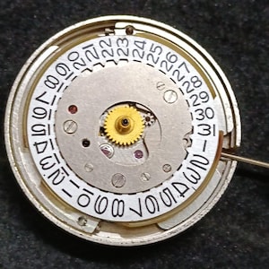 Puede incluir: Un movimiento de reloj mecánico circular con un disco de fecha blanco que tiene los números del 1 al 31 impresos en negro alrededor del borde. Un engranaje central dorado se asienta sobre la placa de metal plateado y sobresale una tija.