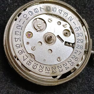 May include: A circular silver-toned mechanical watch movement featuring a date wheel with black printed numbers from 1 to 31. The central plate displays metallic gears, small screws, and a single visible ruby jewel. A small winding crown is attached.
