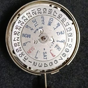 May include: A circular silver-toned watch movement mechanism featuring a white calendar date wheel numbered 1 through 31. The centre plate displays the text SY18-301 and includes day-of-the-week indicators in both English and Arabic script.