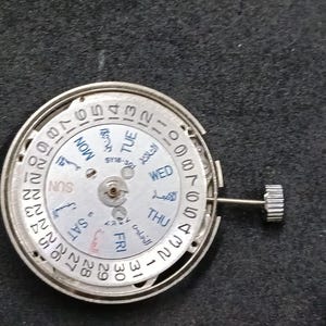 Puede incluir: Un mecanismo de movimiento de reloj circular de tono plateado con una esfera blanca y un anillo de fecha numerado del 1 al 31. La esfera muestra los días de la semana en inglés y árabe, con el texto SY18-301 impreso en el centro y una corona.