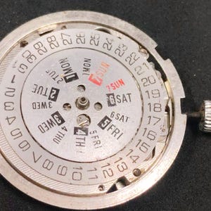 Puede incluir: Un componente de movimiento de reloj circular en tono plateado con un disco de fecha y día. El anillo exterior muestra los números del 1 al 31 en negro. La sección interior muestra los días de la semana en negro, con el domingo en rojo.