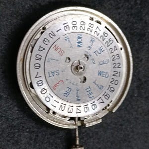 Puede incluir: Un mecanismo de movimiento de reloj circular de tono plateado con un disco de fecha con los números del 1 al 31 y un disco de día que muestra los días de la semana en inglés y árabe, con una pequeña tija de remontuar metálica en la base.