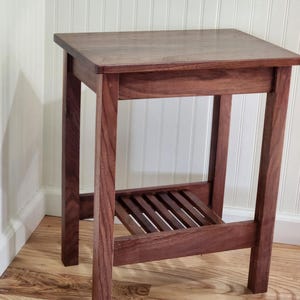 May include: A handcrafted, square-top wooden side table. The table features a dark brown finish, a slatted shelf, and sturdy, square legs. The table measures approximately 24 inches tall.