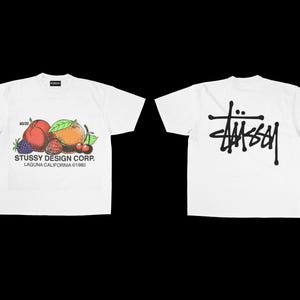 May include: White t-shirts with a fruit graphic and the Stussy Design Corp. logo. One shirt features a colorful illustration of peaches, berries, and cherries. The other shirt displays the Stussy logo in black.