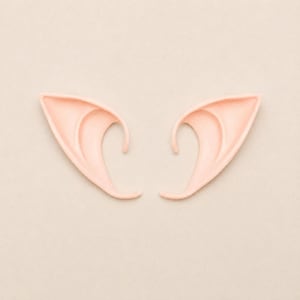May include: A pair of pale peach-colored pointed ear prosthetics. These accessories feature a smooth, curved design with a distinct inner ridge, mimicking the shape of elongated, mythical ears. The surface is matte and uniform in color.