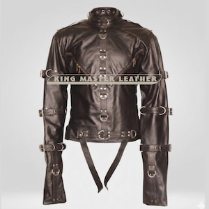 Puede incluir: Chaqueta de cuero marrón oscuro con múltiples correas, hebillas y anillos metálicos. La chaqueta tiene un cuello alto y mangas largas. El texto "KING MASTER LEATHER" está en el pecho. La chaqueta está diseñada con un estilo único.