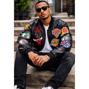 May include: Black leather bomber jacket with multiple team logo patches, including the Houston Rockets, Chicago Bulls, and Los Angeles Lakers. The jacket has a ribbed collar and cuffs, and is worn with a white t-shirt and black jeans.