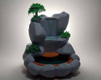 May include: A miniature waterfall fountain with a grey rock formation, a small green tree, and green shrubbery. The water flows into a small orange basin. The fountain is designed to be a decorative item.