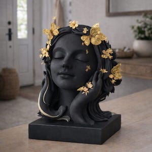 Könnte beinhalten: Schwarze Skulptur eines Frauenkopfes mit geschlossenen Augen, verziert mit goldenen Schmetterlingen und floralen Akzenten. Die Skulptur steht auf einem schwarzen, rechteckigen Sockel, die Hände stützen das Gesicht. Ein elegantes, künstlerisches Dekorationsstück.