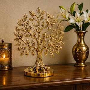 May include: A gold-colored metal tree sculpture with leaves and three birds perched on the branches. The tree sits on a round base. A candle in a decorative lantern and a gold vase with white flowers are also visible.