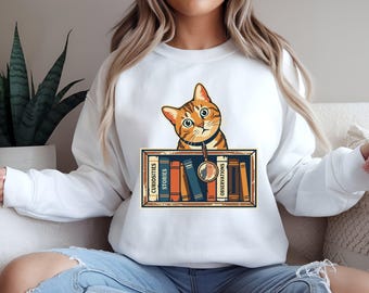 May include: A white crewneck sweatshirt featuring a graphic of an orange tabby cat looking over a bookshelf. The shelf contains colourful books labelled with the words CURIOSITIES, STORIES, and OBSERVATIONS, with a magnifying glass hanging down.