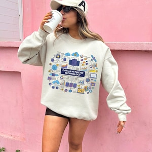 May include: A beige crewneck sweatshirt featuring a colorful graphic print on the chest. The design displays various blue, purple, and yellow icons representing server racks, clouds, and connectivity, with the text DATA & NETWORK TECHNOLOGY.