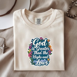 May include: A cream-colored short-sleeve t-shirt featuring a graphic print that reads God is Greater Than the Highs and Lows in white script and block lettering, surrounded by a colorful floral border and small mountain illustrations.
