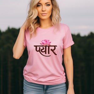 May include: A light pink short-sleeved t-shirt featuring a graphic of a pink lotus flower positioned above the brown Hindi text "प्यार". The shirt has a crew neckline and a standard fit, providing a simple and clean design for casual wear.