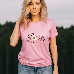 May include: A light pink short-sleeved t-shirt featuring the word Love in a dark pink cursive font across the chest. The letter o is replaced by a white daisy flower with a yellow center. The shirt has a crew neckline and a standard fit.