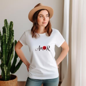 May include: A white short-sleeved t-shirt featuring the word Amor in black script lettering, with a vibrant red flower replacing the letter O. The shirt is paired with a tan fedora hat and blue denim jeans, creating a casual look.