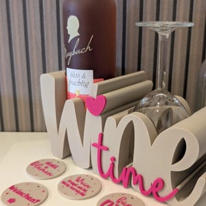 Könnte beinhalten: Ein beigefarbener Wein- und Glashalter mit dem Wort Wine in großen Buchstaben, einem rosa Herz und dem Wort time in rosa Schreibschrift. Vorne liegen vier runde, beige Untersetzer mit rosa deutschem Text und kleinen Punkten.