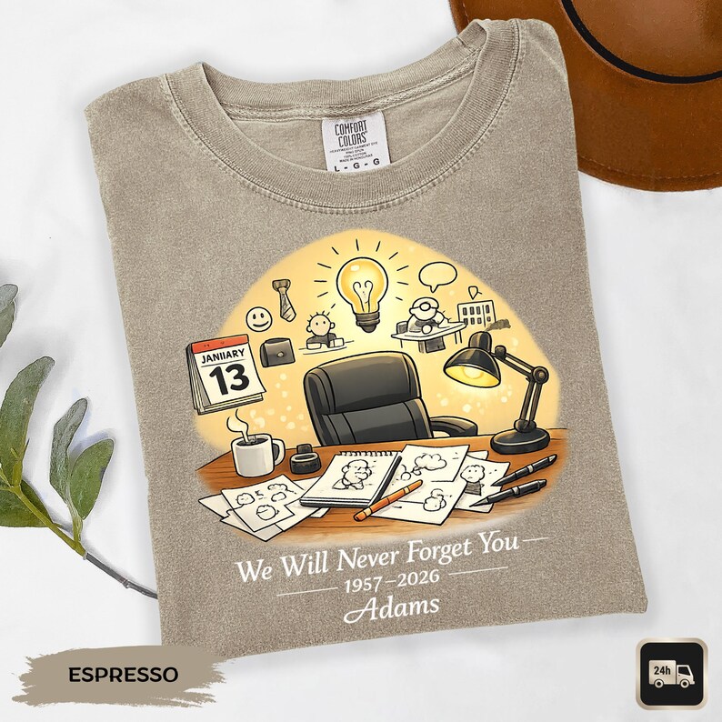 May include: Espresso-colored t-shirt with a graphic design featuring a desk scene with a calendar, coffee cup, and drawings. The text reads "We Will Never Forget You" and "Adams".