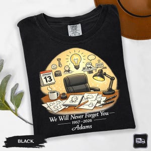 May include: Black t-shirt with a graphic design featuring a desk scene with a calendar, coffee cup, and drawings. The text reads "We Will Never Forget You - 1957-2026 Adams."