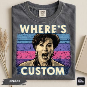 May include: Pepper-colored t-shirt with a graphic featuring a woman's face and the words "WHERE'S CUSTOM" in a retro font. The design includes blue, purple, and pink horizontal stripes. The shirt has a crew neck and a Comfort Colors tag.