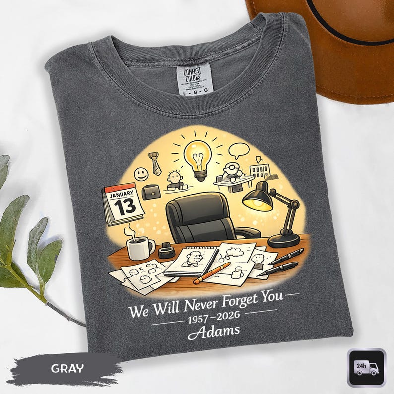 May include: Gray Comfort Colors t-shirt with a graphic design featuring a desk scene with a calendar, coffee cup, and drawings. The text reads "We Will Never Forget You - 1957-2026 Adams."