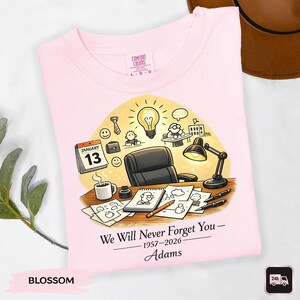 May include: A light pink t-shirt with a graphic design featuring a desk scene with a calendar, coffee cup, and drawings. The text reads "We Will Never Forget You - 1957-2026 - Adams". The shirt is labeled "Blossom".