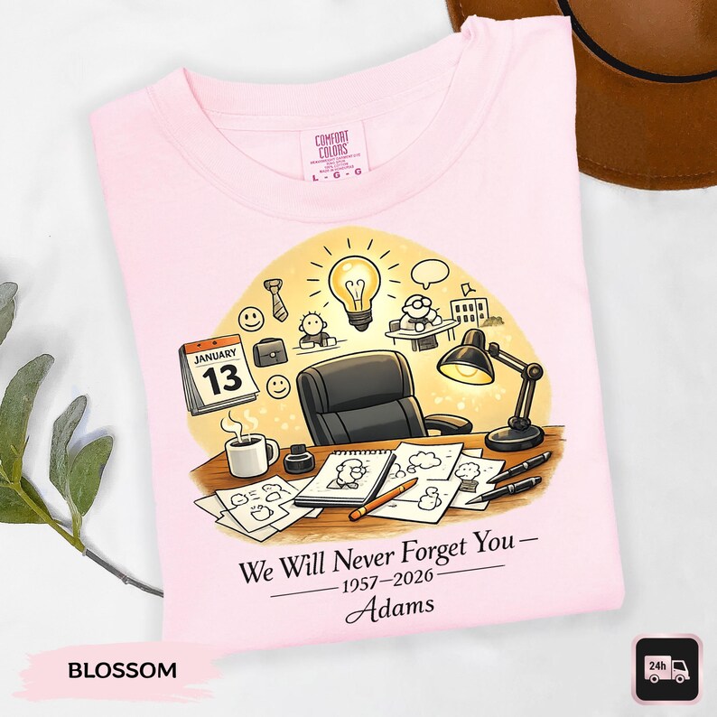 May include: A light pink t-shirt with a graphic design featuring a desk scene with a calendar, coffee cup, and drawings. The text reads "We Will Never Forget You - 1957-2026 - Adams". The shirt is labeled "Blossom".