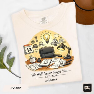 May include: Ivory-colored t-shirt featuring a desk scene with a calendar showing January 13th, a lightbulb, and the text "We Will Never Forget You - 1957-2026 - Adams." The shirt has a Comfort Colors label.