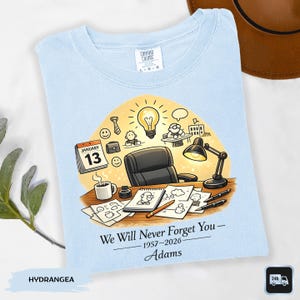May include: Light blue Comfort Colors t-shirt with a graphic design. The design features a desk scene with a calendar, coffee cup, and the text "We Will Never Forget You - 1957-2026 - Adams".