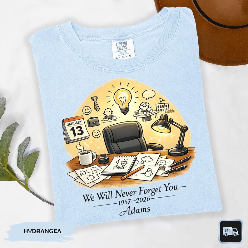 May include: Light blue Comfort Colors t-shirt with a graphic design. The design features a desk scene with a calendar, coffee cup, and the text "We Will Never Forget You - 1957-2026 - Adams".