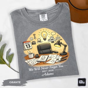 May include: Gray t-shirt with a graphic of an office scene, including a desk, lamp, and calendar. The calendar shows January 13. Text on the shirt reads "We Will Never Forget You" and the name "Adams" with dates. The shirt has a Comfort Colors tag.