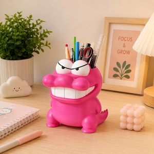 May include: A bright pink alligator-shaped desk organizer with white teeth and eyes. The organizer holds pens, pencils, and a ruler. A framed print with the words "FOCUS & GROW" is in the background.