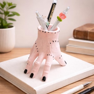May include: A pink hand-shaped pen holder with black nail polish and stitched details. The holder contains several pens and pencils. The holder is resting on a white book. A unique desk accessory.