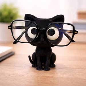 May include: A black cat-shaped eyeglass holder with large white eyes and a pair of black-framed eyeglasses. The holder is made of a glossy material and sits on a wooden surface. The cat is in a sitting position.
