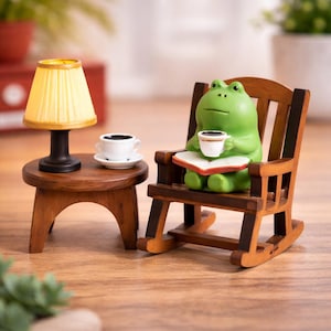 May include: A charming miniature scene with a green frog figurine in a wooden rocking chair, holding a coffee cup and reading. A small wooden side table with a lamp and a coffee cup and saucer add to the cosy atmosphere.