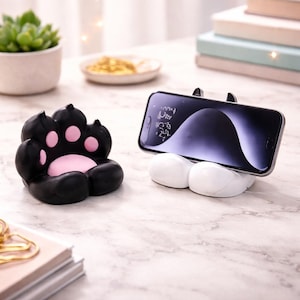 Peut inclure: Un support de téléphone en forme de patte noire et rose et un support de téléphone blanc en forme de pattes de chat. Un smartphone à écran sombre est posé sur le support. Sur une surface en marbre.