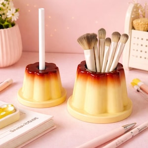 Puede incluir: Dos soportes con forma de flan, uno con un palito blanco y otro con brochas de maquillaje. Los soportes son amarillos con la parte superior marrón, parecidos a un postre. Se encuentran sobre una superficie rosa claro.