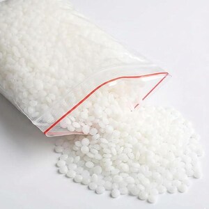 May include: A clear plastic bag with a red zipper is partially open, spilling out a pile of small, white, rounded beads. The beads are scattered on a white surface. The bag is filled with the same white beads.