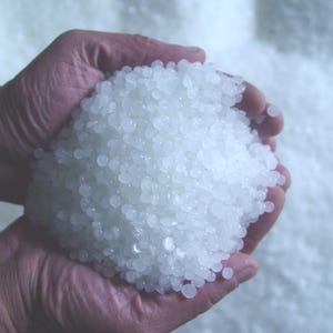 May include: A close-up of hands holding a large pile of small, translucent, white, round beads. The beads are tightly packed together, filling the hands and spilling over the sides. The background is a soft, out-of-focus white.