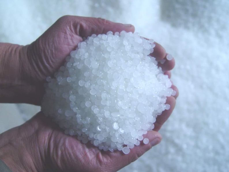 May include: A close-up of hands holding a large pile of small, translucent, white, round beads. The beads are tightly packed together, filling the hands and spilling over the sides. The background is a soft, out-of-focus white.