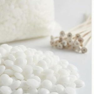 May include: A close-up shot of white candle-making supplies. A pile of white wax beads is in the foreground, with a bag of wax beads and a bundle of wicks in the background. The items are on a white surface.