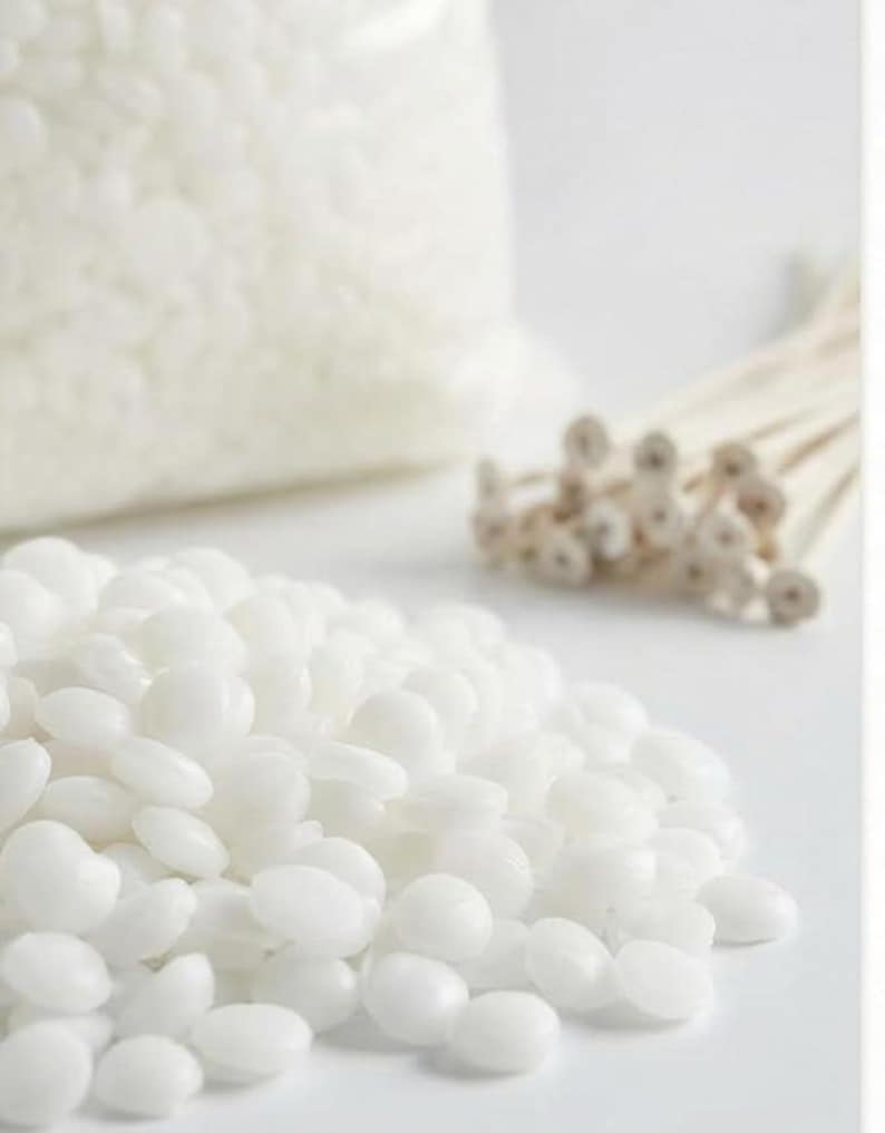 May include: A close-up shot of white candle-making supplies. A pile of white wax beads is in the foreground, with a bag of wax beads and a bundle of wicks in the background. The items are on a white surface.