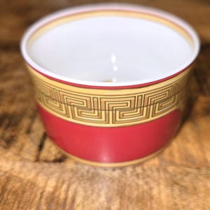 Puede incluir: Un cuenco peque&ntilde;o y redondo de cer&aacute;mica con una base de color rojo vibrante y un interior blanco. La parte superior exterior est&aacute; decorada con un patr&oacute;n de greca en negro y dorado, bordeado por finas l&iacute;neas doradas.