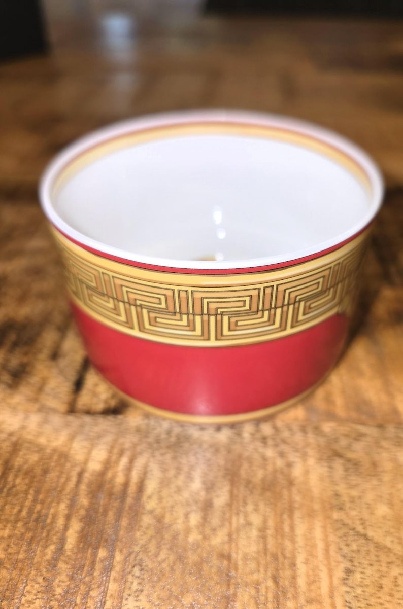 Puede incluir: Un cuenco peque&ntilde;o y redondo de cer&aacute;mica con una base de color rojo vibrante y un interior blanco. La parte superior exterior est&aacute; decorada con un patr&oacute;n de greca en negro y dorado, bordeado por finas l&iacute;neas doradas.