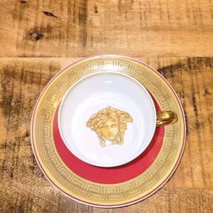 Puede incluir: Un juego de taza y plato de porcelana blanca con un emblema de cabeza de Medusa dorada en el centro de la taza. El plato tiene una banda roja ancha bordeada por un patr&oacute;n de llave griega dorada. La taza incluye un asa dorada y un borde a juego.