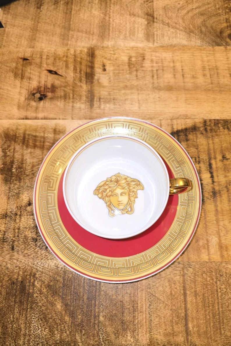 Puede incluir: Un juego de taza y plato de porcelana blanca con un emblema de cabeza de Medusa dorada en el centro de la taza. El plato tiene una banda roja ancha bordeada por un patr&oacute;n de llave griega dorada. La taza incluye un asa dorada y un borde a juego.